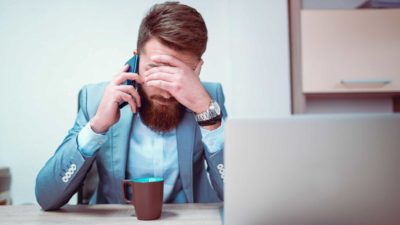 Hipster man puts head in hand as he talks on phone in front while sitting at a desk.