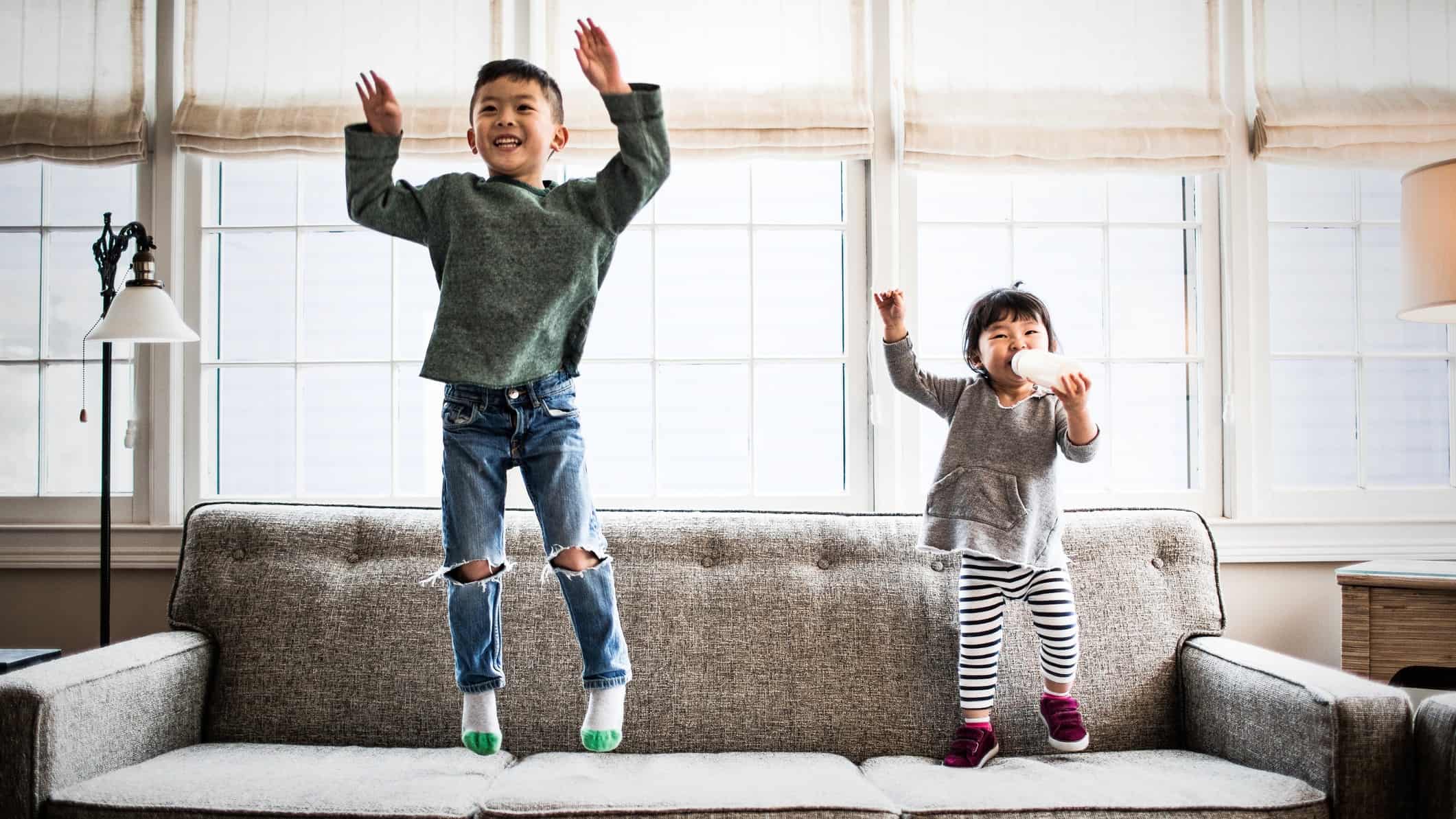 A young boy and young girl jump on the sofa with their hands in the air.