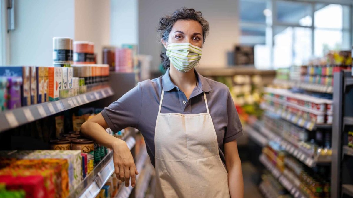 Female shop attendant wearing apron and mask standing in grocery aisle in small local shop