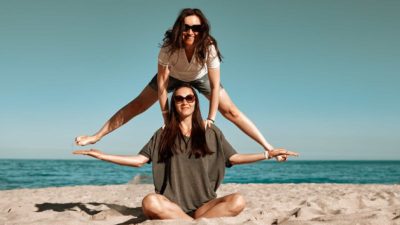 Woman sits in lotus position on the sand in front of a lake as another woman leapfrogs over her.