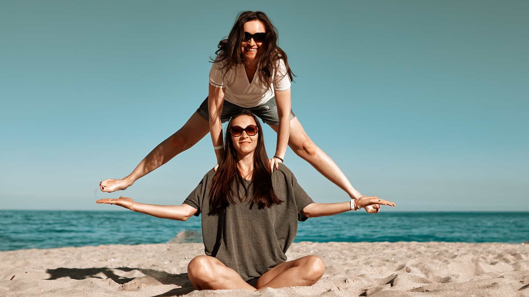 Woman sits in lotus position on the sand as another woman leapfrogs over her.