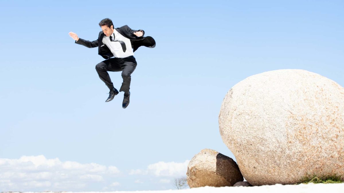 Man in a business suit leaps off a boulder in front of a blue sky.