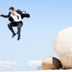 Man in a business suit leaps off a boulder in front of a blue sky.