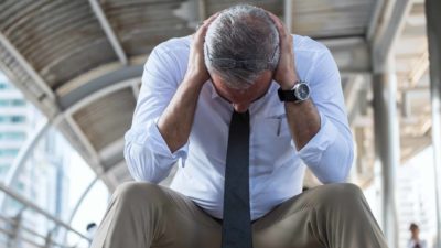 Older businessman sits slumped with head down and hands on either side of his head.