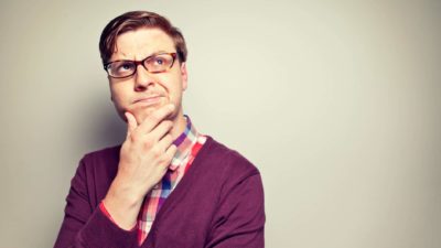 A man wearing glasses and a purple vest holds his hand to his chin and wonders