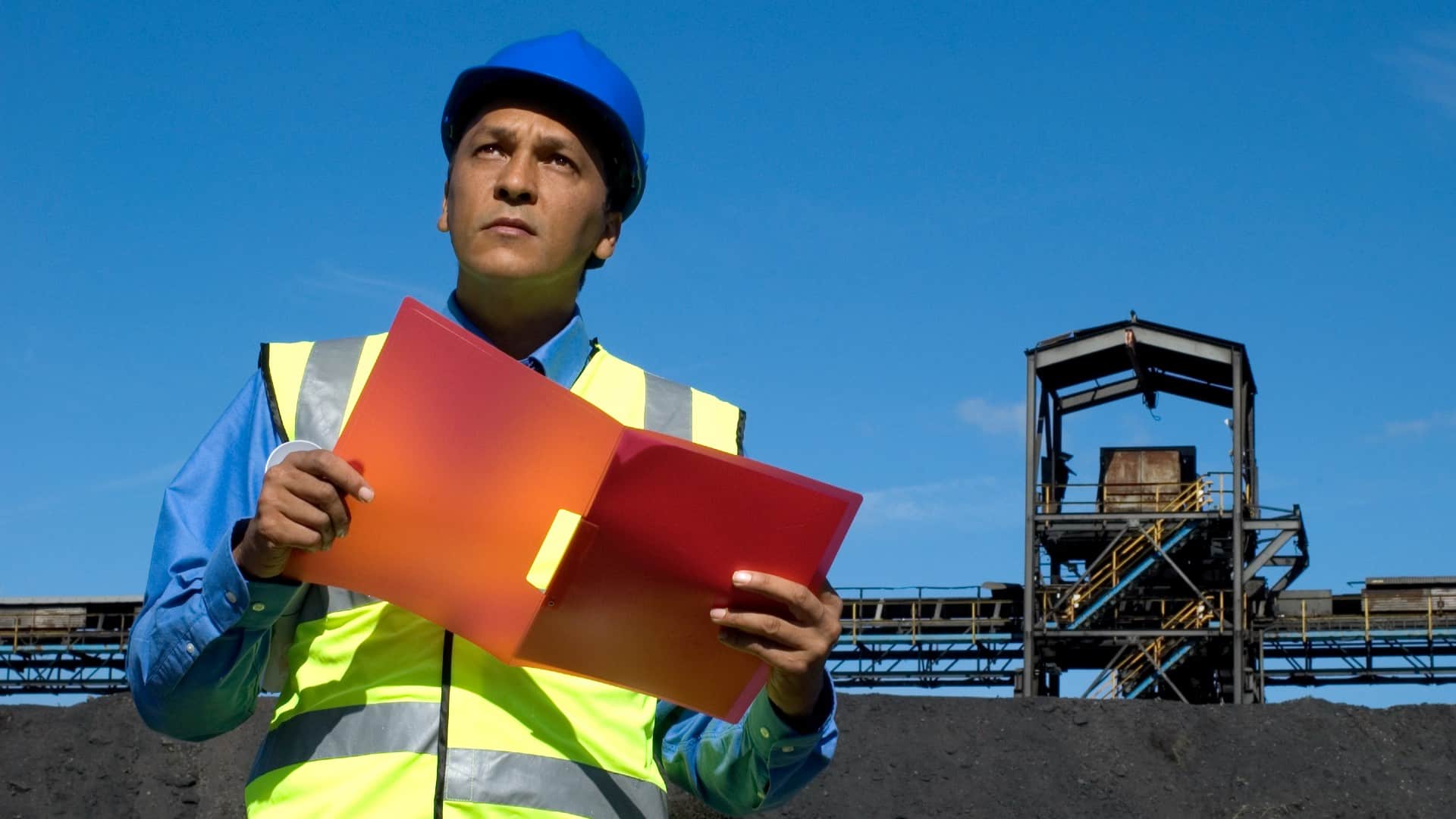 Australian Strategic Materials employee wearing a hard hat at a mine looks into the distance as he checks a folder.