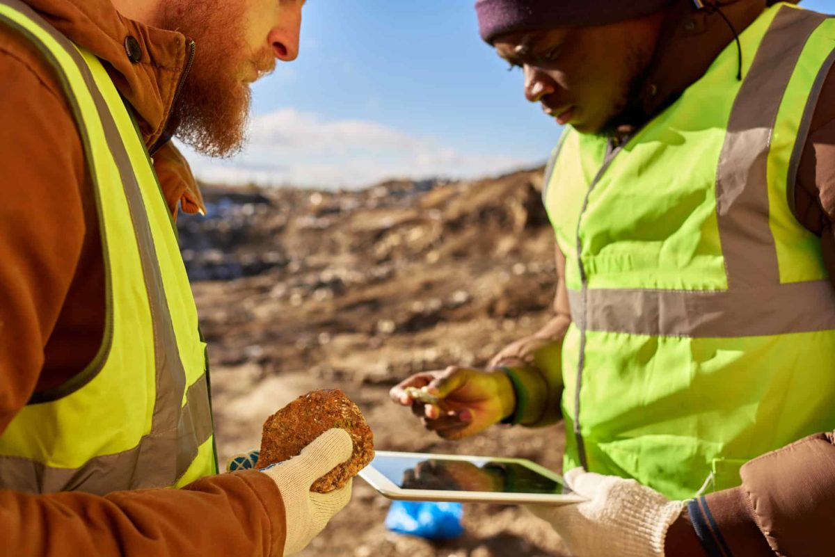 Two miners dressed in hard hats and high vis gear standing at an outdoor mining site discussing a mineral find with one holding a rock and the other looking at a tablet.