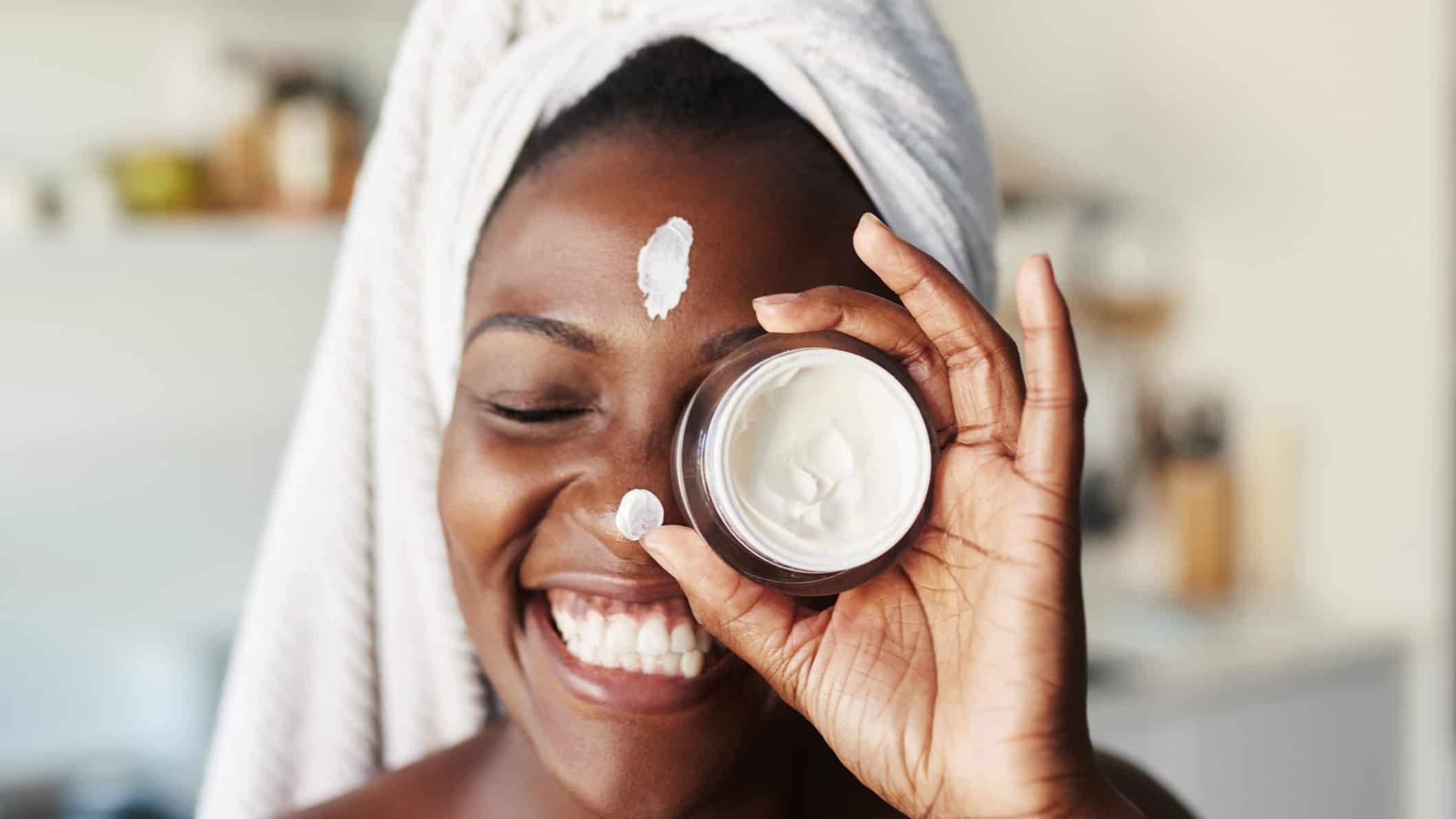 smiling woman with towel on head holds pot of face cream in front of eye