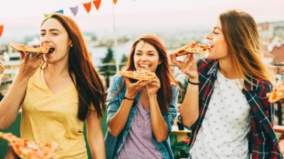 Three women smile and laugh as they eat pizza at a rooftop party.