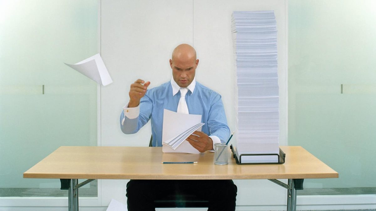 A man in a suit at a desk throws papers around onto the floor as he reads them.