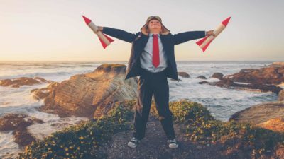 A boy stands firm on a rocky cliff holding a rocket in each hand and looking up toward the sky, anticipating flying into space.