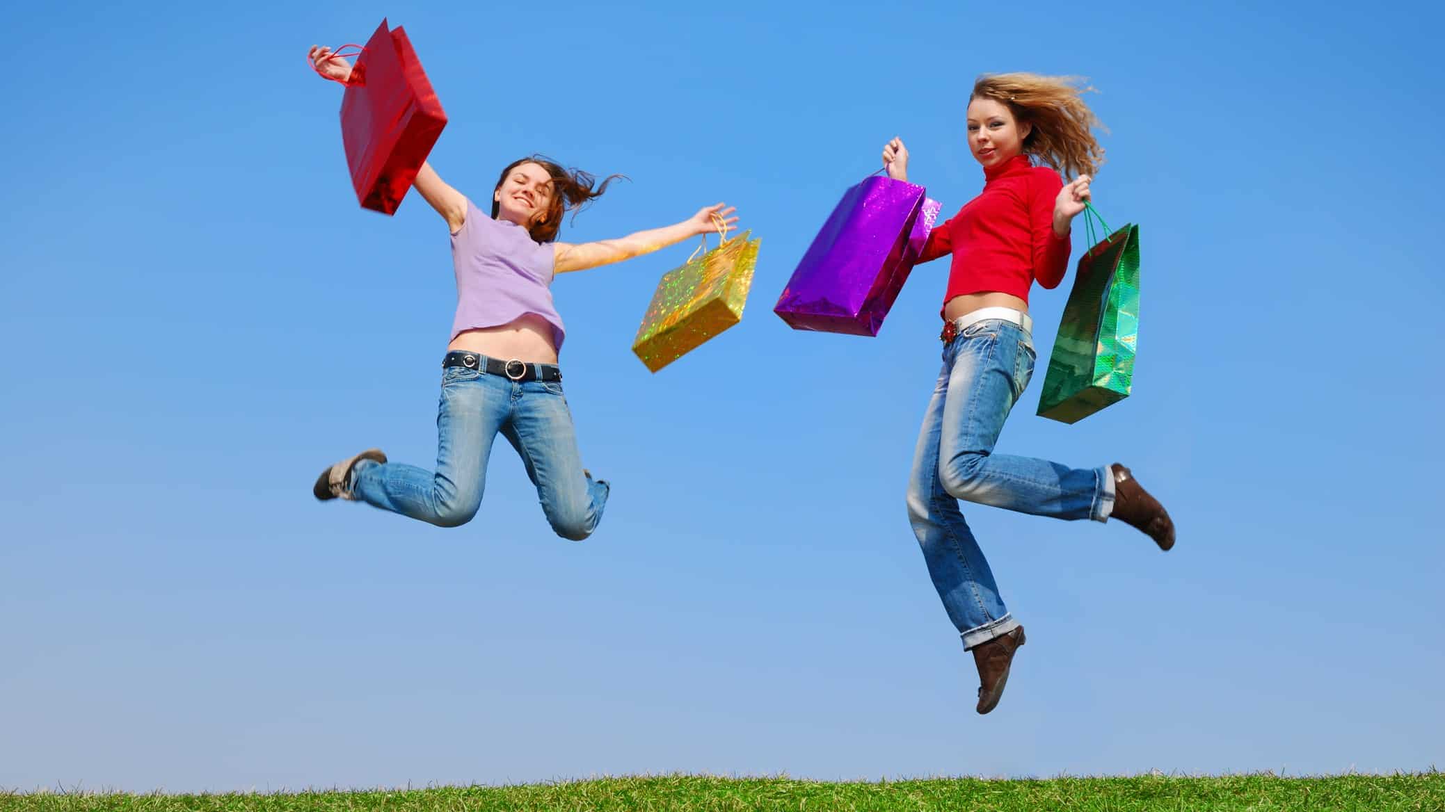 Two women with shopping bags leap on the grass in front of a blue sky.