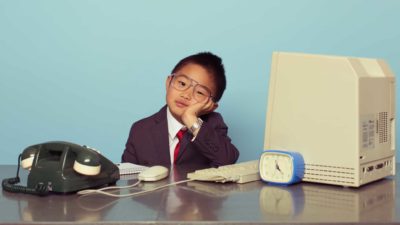 A boy in a business suit sits at a retro desk with old phone and computer, indicating a slowdown in bank shares