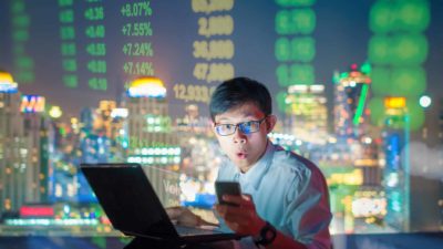 Man looks shocked as he works on laptop on top a skyscraper with stockmarket figures in graphic behind him.