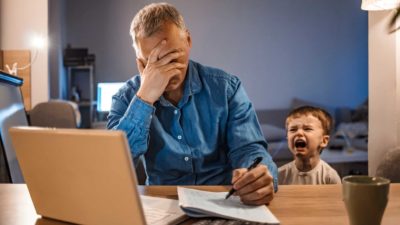 A stressed man sits with head in hands at laptop as small child cries next to him.