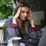 Young woman dressed in suit sitting at cafe staring at laptop screen with hands to her forehead looking tense.