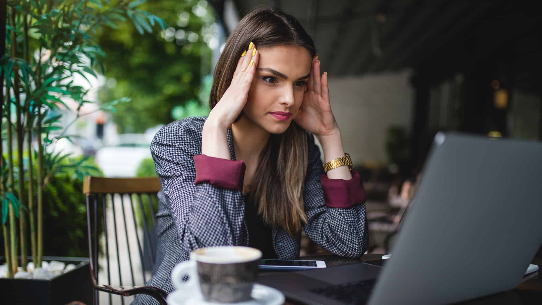 Young woman dressed in suit sitting at cafe staring at laptop screen with hands to her forehead looking tense