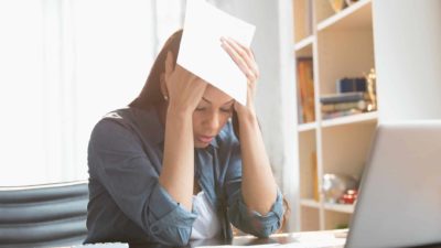 Woman puts head in hands as she sits at her computer trying to pay bills.