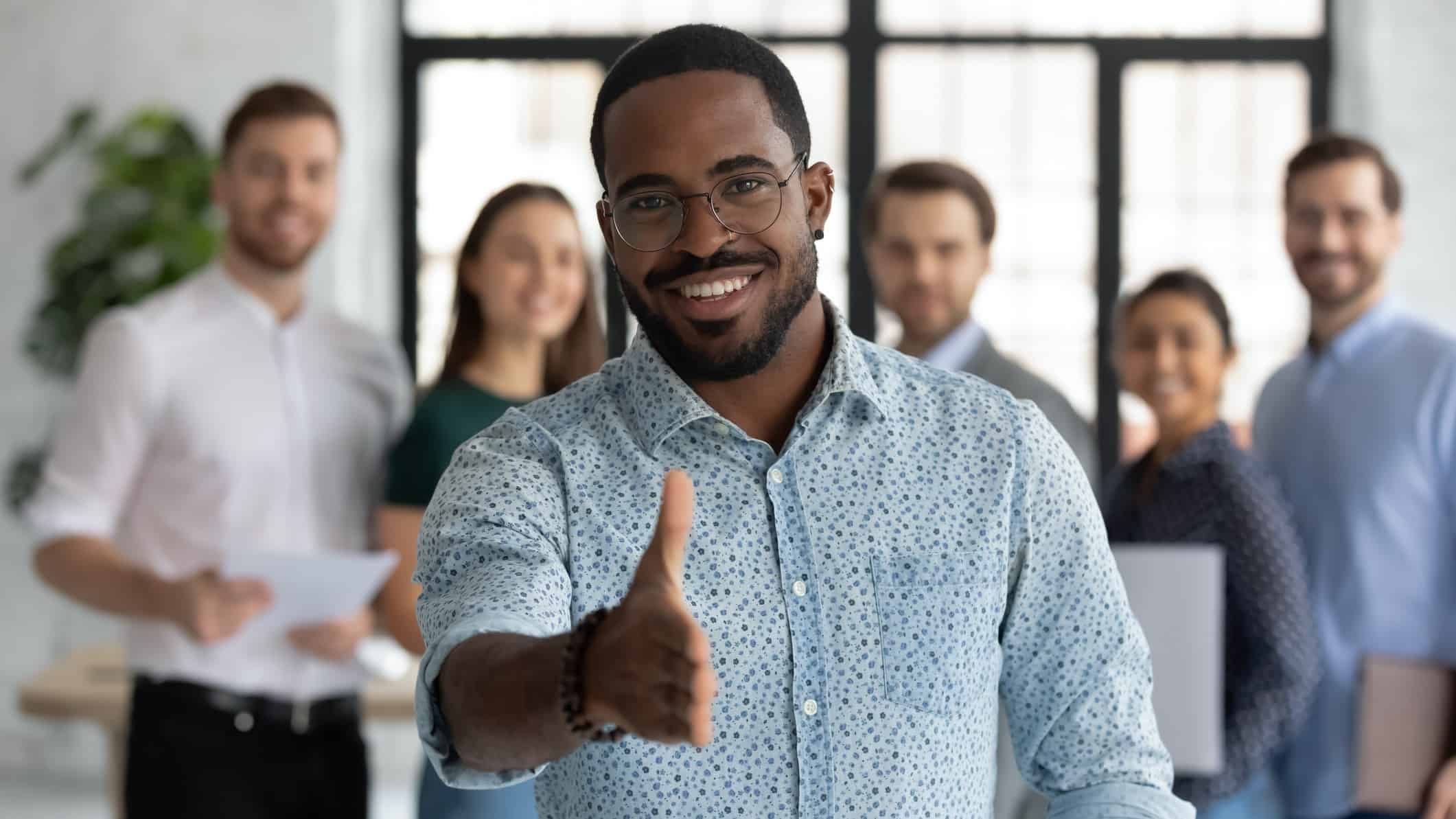A man standing in front of co-workers extends his hand in welcome