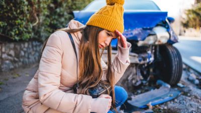 Frustrated woman crouches next to wrecked car after a car crash feeling shocked by glad she has QBE insurance