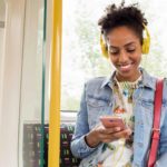 A woman smiles as she sits on the bus using her phone and listening to music through headphones.