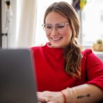A young woman wearing glasses and a red top looks at her laptop smiling