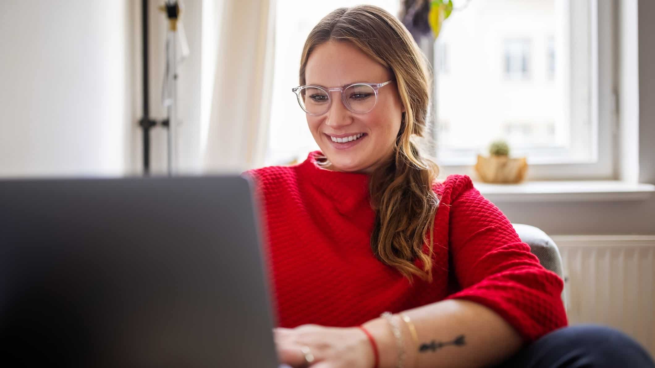 A young woman wearing glasses and a red top looks at her laptop happily, watching, waiting, on good results.