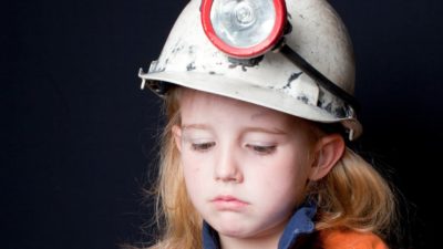 Young girl wearing a hard hat and light looks downcast.