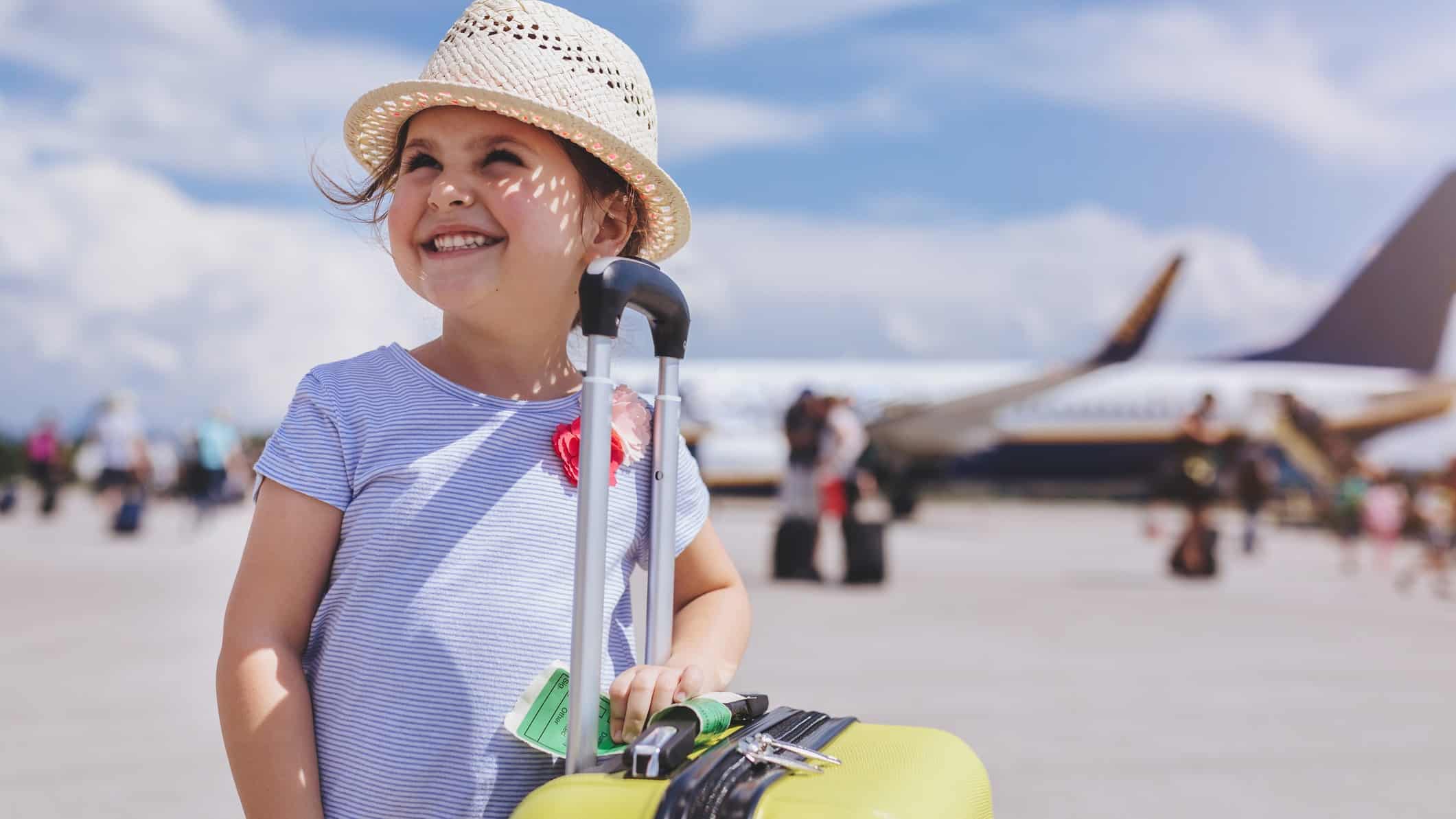 Young girl smiles with her hand on top of a suitcase while standing on the tarmac with an aeroplane in the background.