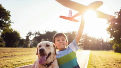 A boy hugs his dog with one arm and holds a big red plane in the air with the other in the beautiful sunshine.
