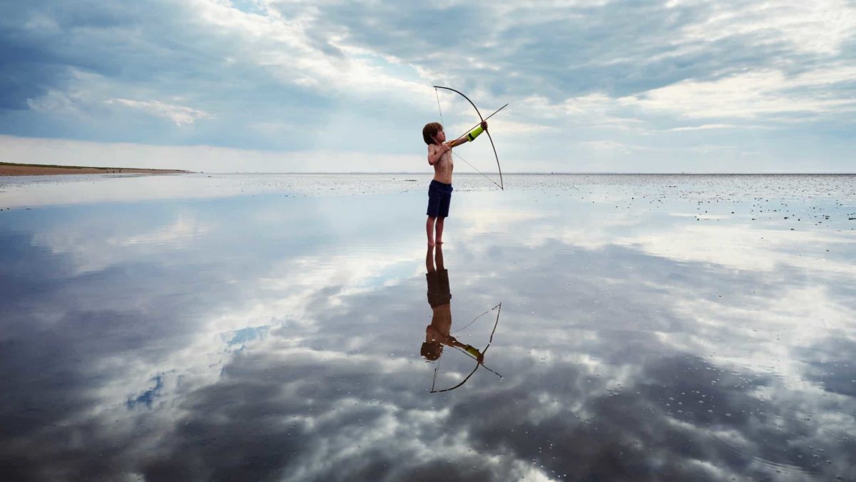A boy stands in still ankle-deep water brandishing a bow and arrow.