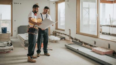Two construction workers stand in a half-finished apartment looking at blueprints together