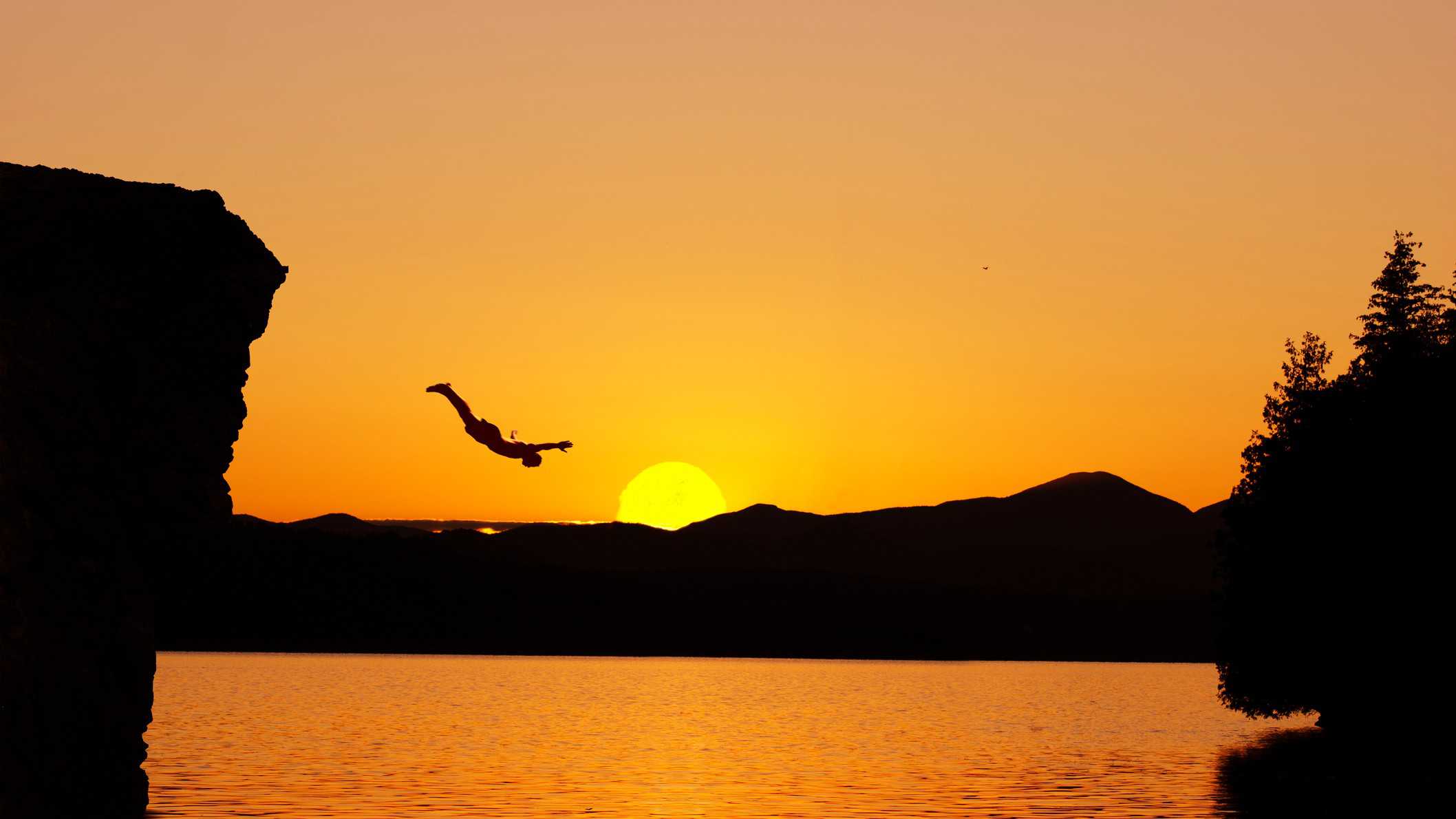 A person dives from a cliff into the water with a golden sunset in the background.