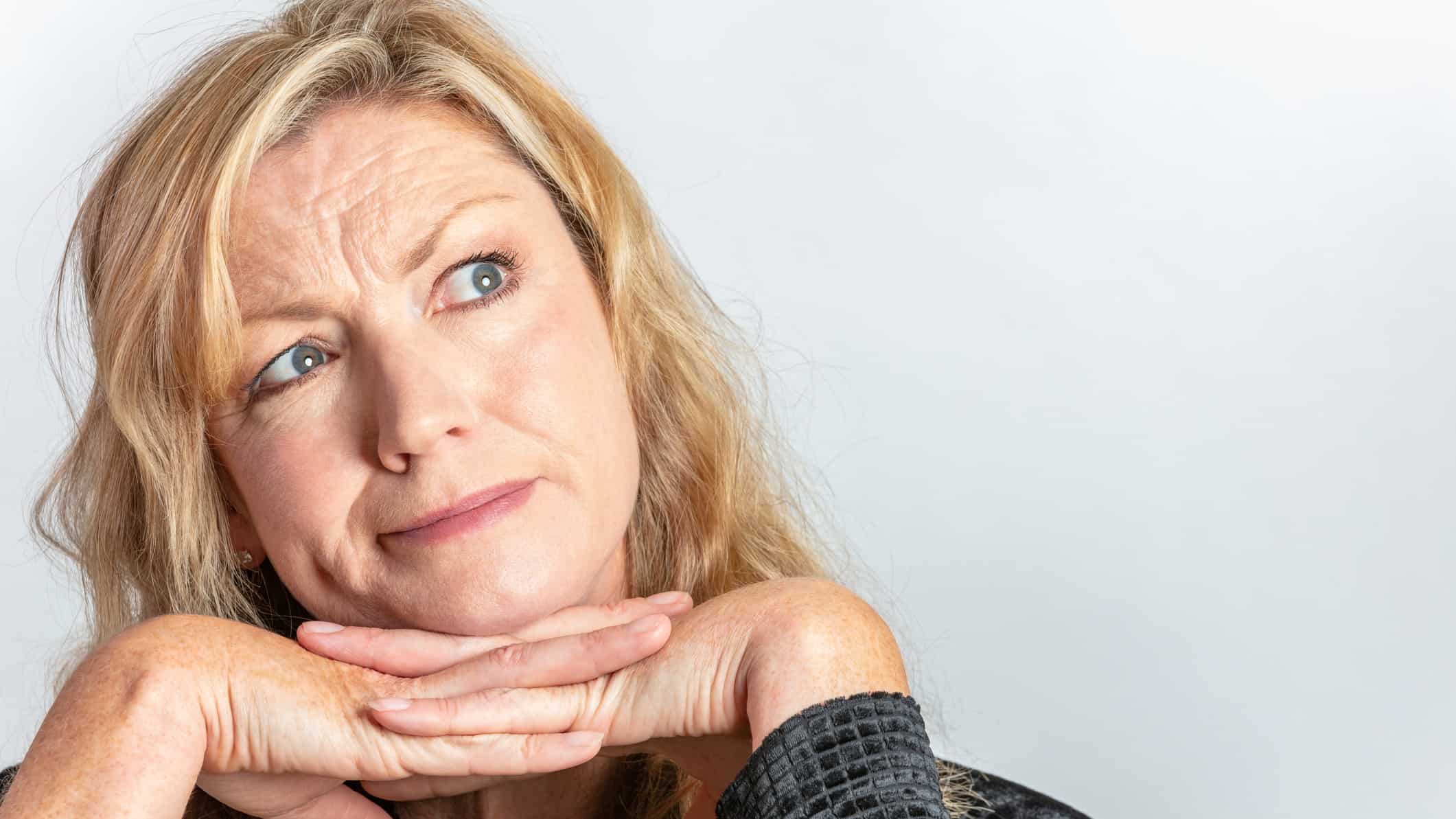 a woman frowns slightly and looks with her eyes to the side as though she is pondering a question. She holds her chin resting on both hands.