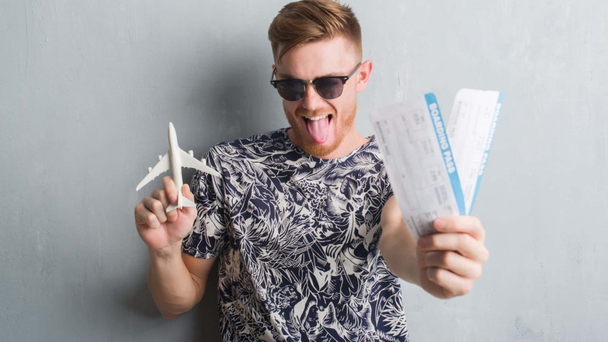 a man in a flowery t-shirt and sunglasses clutches two airline boarding passes and a toy plane in his other hand and smiles widely to the camera, sticking his tongue out to show his joy.