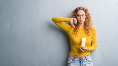 A woman wearing a gold top and carrying a gold bar gives the thumbs down signal as she leans against a wall with a sombre look on her face as the Kingsgate share price goes lower