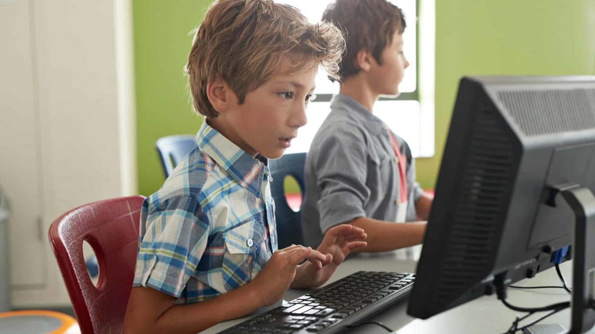 children sit side by side at respective computer screens, both deep in concentration as though they are doing online lessons.