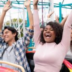 two people sit side by side on a rollercoaster ride with their hands raised in the air and happy smiles on their faces