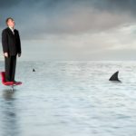 A man in a business suit stands on top of an office chair in a sea of murky water with shark fins circling.