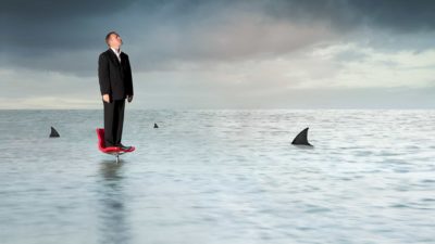a man in a business suit stands on top of an office chair in a sea of murky water with shark fins circling.
