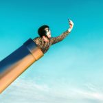 a man wearing old fashioned aviator cap and goggles emerges from the top of a cannon pointed towards the sky. He is holding a phone and taking a selfie.
