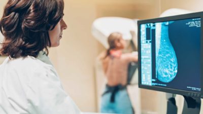 a sonographer monitors an image of a patient's breast on a screen with the patient standing at an imaging device in the background.