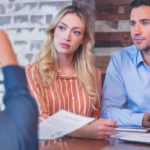 a couple consider the advice from a man with documents laid out on a table and the man holding a tablet in his hand.
