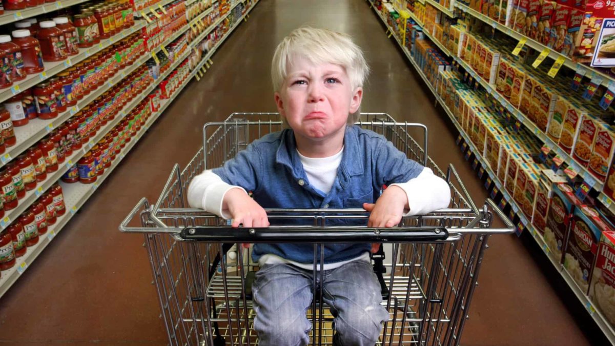 A child pulls a very sad crying face sitting in the child seat of a supermarket trolley in a supermarket aisle lined with grocery items.