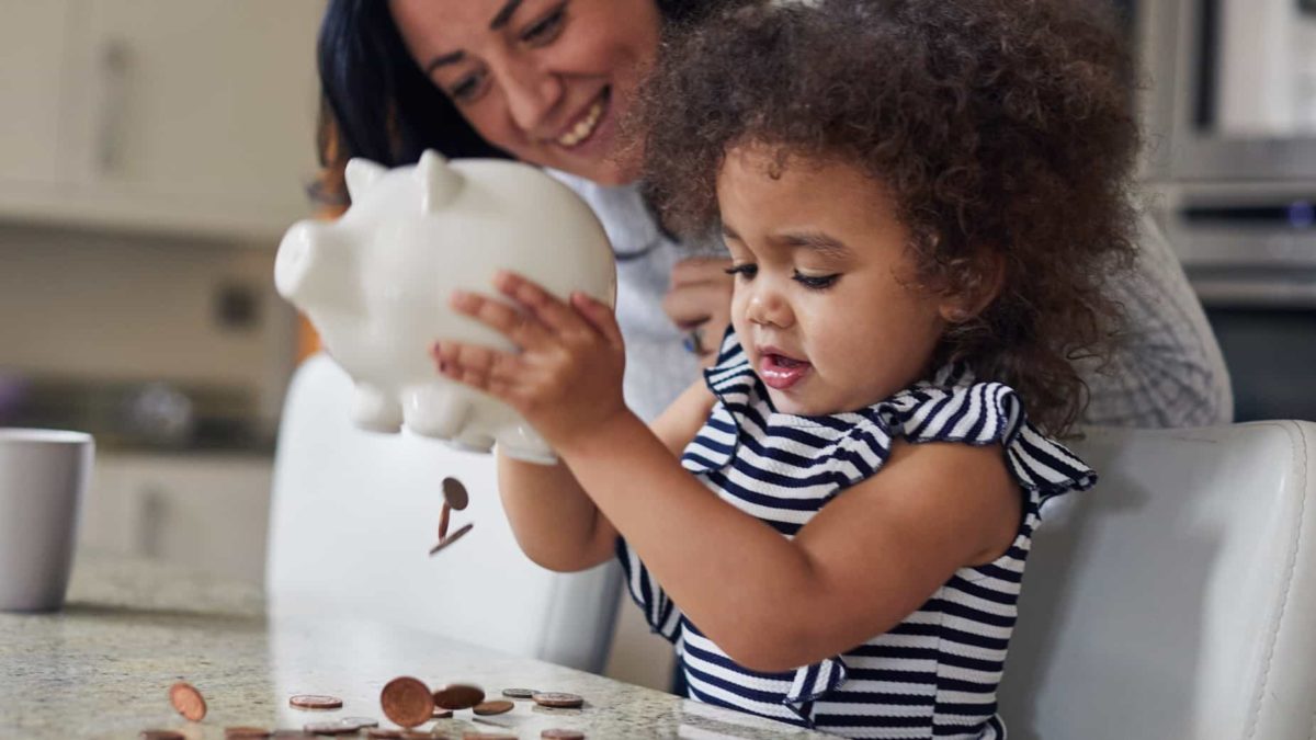 a small girl empties a piggy bank of coins onto a table while her mother looks on in the background.