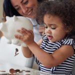A small girl empties a piggy bank of coins onto a table while her mother looks on in the background.