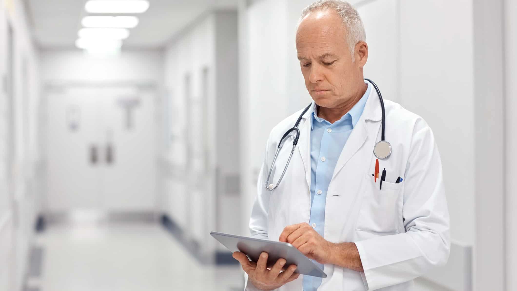a doctor in a white coat with a stethoscope around his neck stands in the hallway of a hospital deep in concentration over a tablet device in his hands.