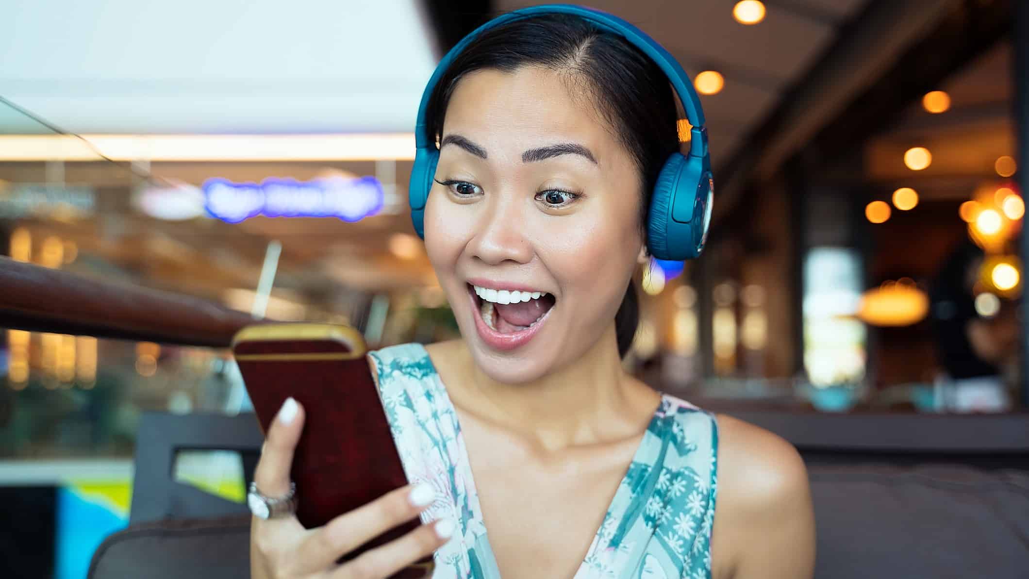 A woman wearing headphones looks delighted and animated on news she's receiving from her mobile phone that she is holding close to her face.