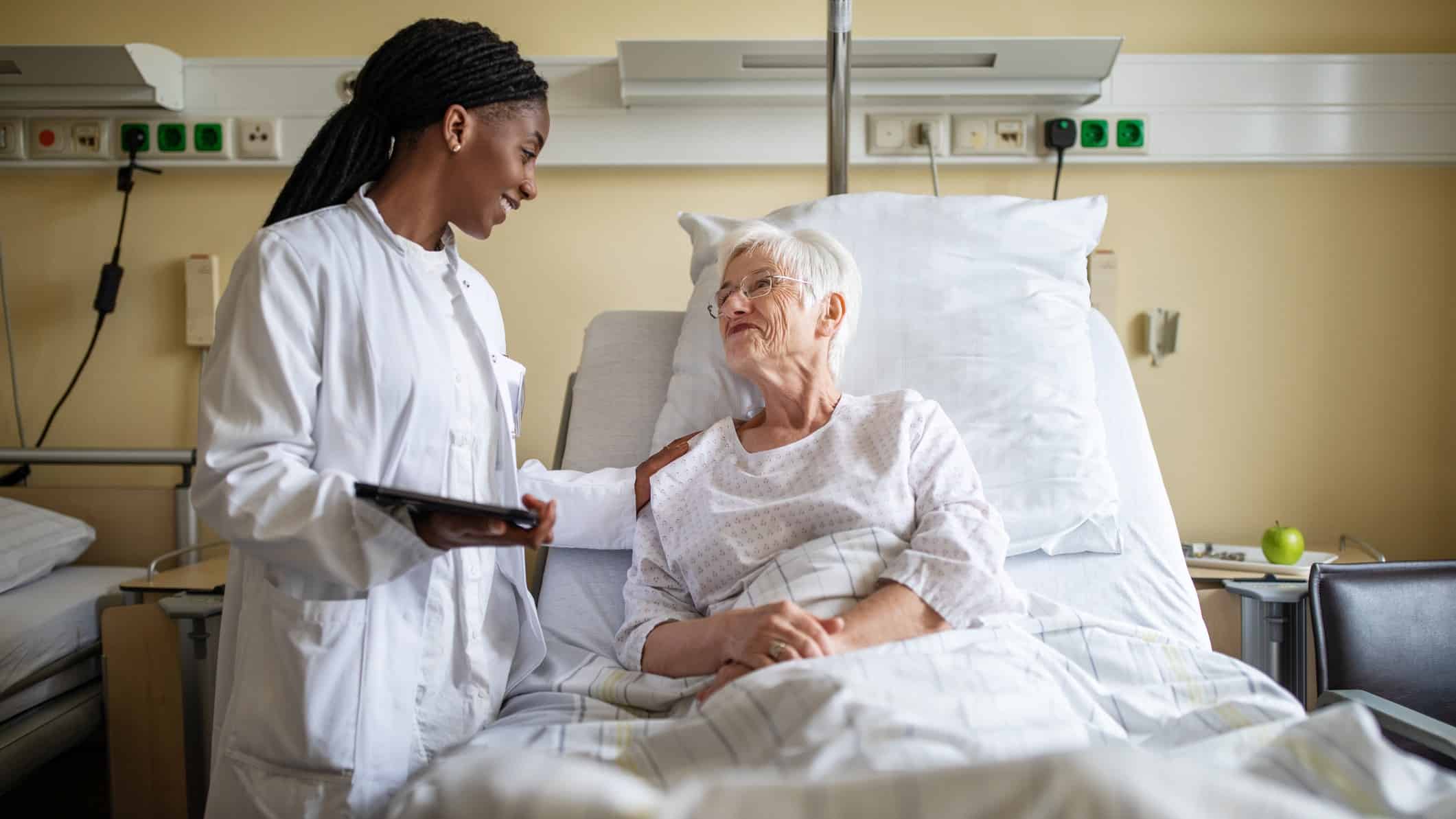a patient in a hospital bed is reassured by a doctor at her bedside with the doctor placing a hand on the paient's shoulder. They are both smiling.