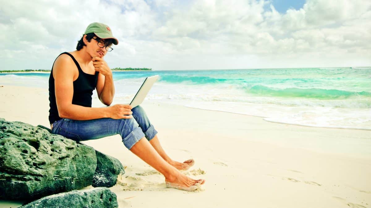 a man sits on a beach on a rock looking at a laptop computer with a puzzled and disappointed look on his face and his hand to his chin.